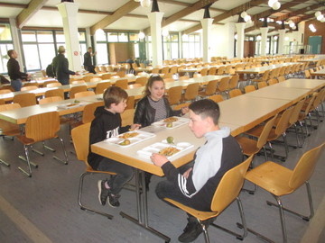 drei Kinder sitzen in einer riesigen Halle voller Tische und Stühlen vor Essentabletts.